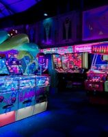 An indoor arcade filled with vibrant gaming machines lit with neon blue and pink lights. The scene includes various arcade games with flashing screens and colorful displays, and a large overhead structure resembling a green snake. The background shows illuminated signs and themed decor, contributing to an immersive entertainment atmosphere.
