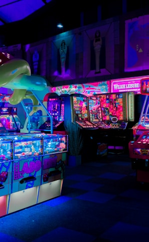 An indoor arcade filled with vibrant gaming machines lit with neon blue and pink lights. The scene includes various arcade games with flashing screens and colorful displays, and a large overhead structure resembling a green snake. The background shows illuminated signs and themed decor, contributing to an immersive entertainment atmosphere.