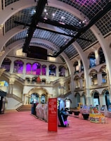 The interior of a large, ornately designed building with arched ceilings and intricate architectural details. The space features gaming machines lined up along the floor, under a high ceiling with a grid pattern. Lighting in purple and white tones enhances the ambiance.