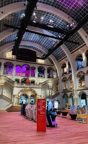 The interior of a large, ornately designed building with arched ceilings and intricate architectural details. The space features gaming machines lined up along the floor, under a high ceiling with a grid pattern. Lighting in purple and white tones enhances the ambiance.
