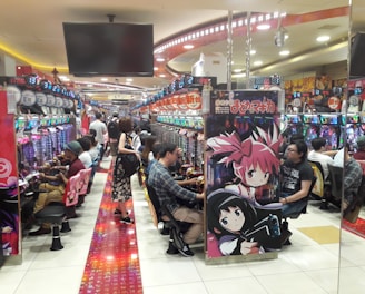 A bustling arcade filled with people playing various gaming machines. Brightly colored displays and animated characters are prominent on posters and screens. The atmosphere is lively with patrons seated in rows, focusing on the games. Overhead signage and electronic scoreboards are visible, contributing to the busy and vibrant environment.