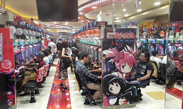A bustling arcade filled with people playing various gaming machines. Brightly colored displays and animated characters are prominent on posters and screens. The atmosphere is lively with patrons seated in rows, focusing on the games. Overhead signage and electronic scoreboards are visible, contributing to the busy and vibrant environment.