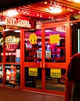 A brightly lit entrance to a casino with neon signs, including one advertising a £500 jackpot. The doors have clear signage indicating entry is restricted to those over 18. A person in a red jacket stands to the right, possibly interacting with a machine or display.