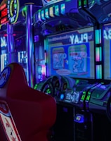 A vibrant arcade environment with several illuminated gaming machines. The image features arcade racing games with visible steering wheels, colorful control panels, and screens displaying game instructions. Bright LED lights in various colors illuminate the machines and surrounding area.