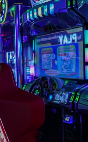 A vibrant arcade environment with several illuminated gaming machines. The image features arcade racing games with visible steering wheels, colorful control panels, and screens displaying game instructions. Bright LED lights in various colors illuminate the machines and surrounding area.